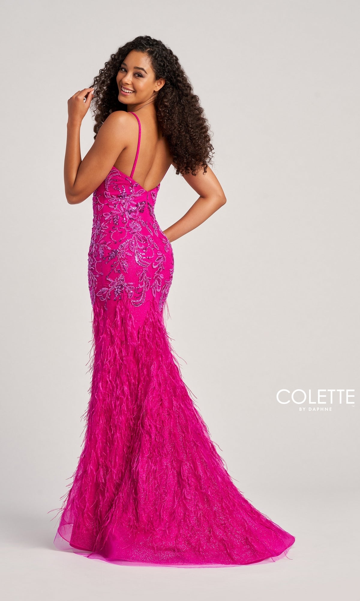 A woman poses and smiles in the Long Colette CL5103 Feathered Mermaid Prom Dress, a bright pink, floor-length gown with intricate beading, feather accents, and a backless design against a plain background.